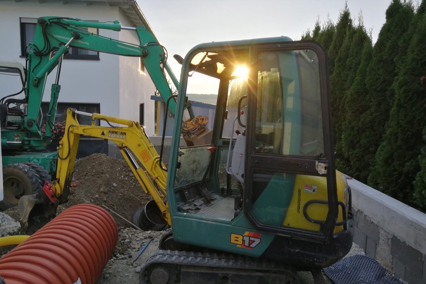 Minibagger im Gegenlicht neben orangen Drainagerohren